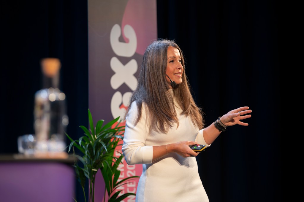 Kvinna med talarmick på huvudet, utsläppt hår. I ena handen en klicker till datorn den andra gestikulerar med spretiga fingrar. Blicken är bortåt. Vit klänning. Skylt i bakgrunden med bokstäverna SXC.